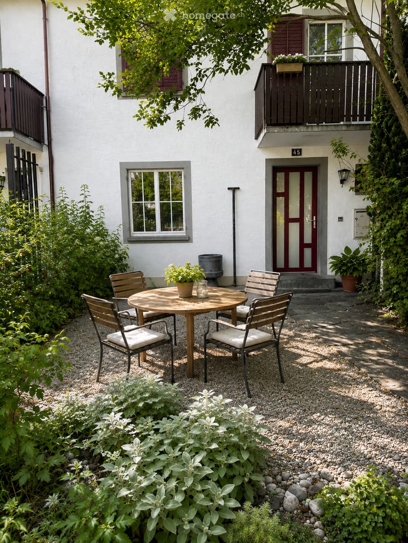 A small, well-maintained garden area with a round wooden table and four matching chairs, surrounded by greenery and a gravel pathway.
