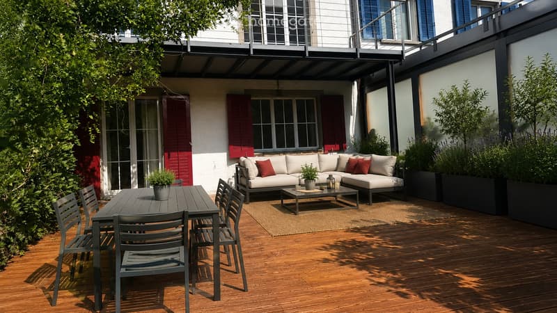 A modern outdoor patio with wooden flooring, a grey sofa with red cushions, a coffee table, a dining table with chairs, and potted plants.