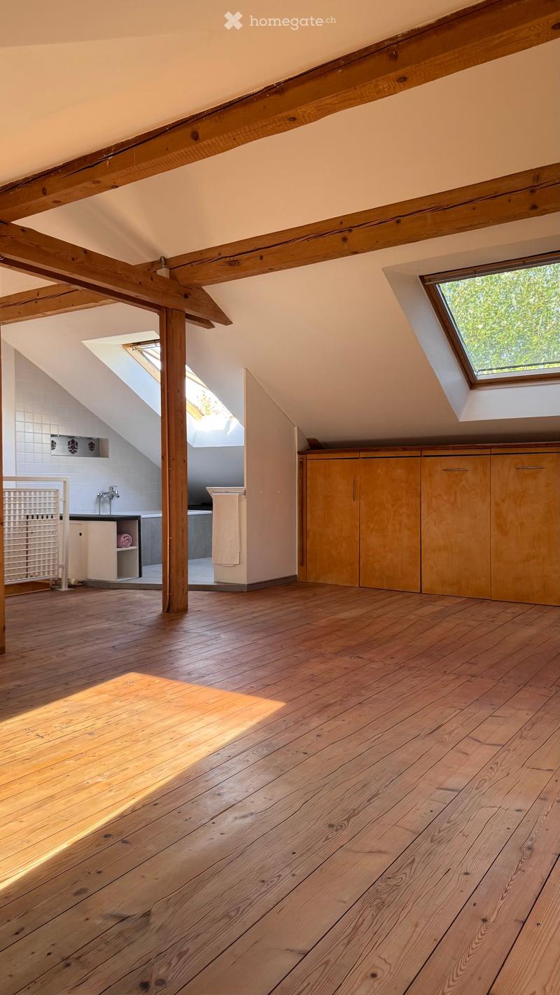Bright, open attic space with wooden beams, wooden flooring, skylights, and built-in wooden cabinets. A small bathroom is visible in the background.