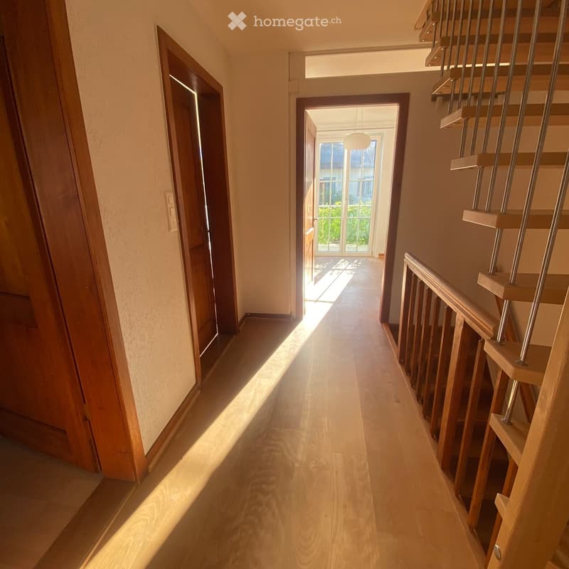 Bright hallway with wooden flooring, modern staircase with metal railings, and open doors leading to other rooms.