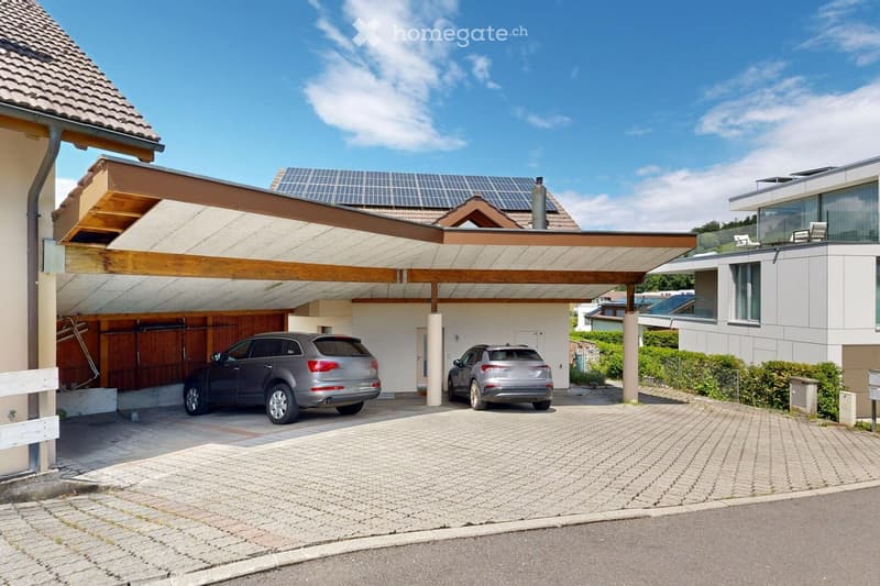 Two-story house with a covered driveway, two parked cars, solar panels on the roof, and a modern design with large windows and a balcony area.