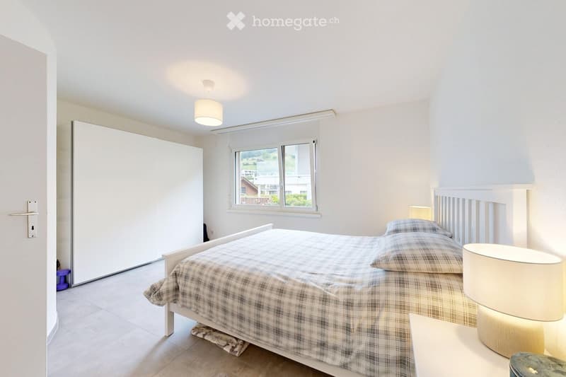 Bright bedroom with a double bed, plaid duvet, nightstand with lamp, and a large window with blinds. The room features a white wardrobe and a modern ceiling light.