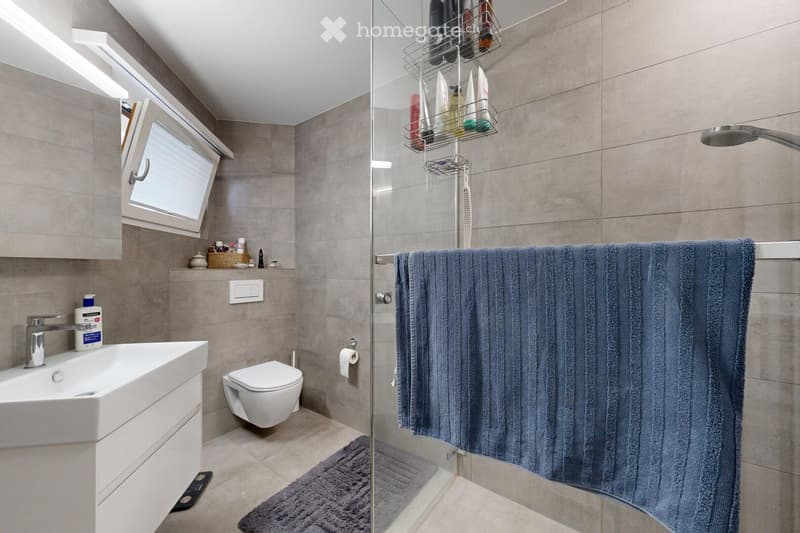 Modern bathroom with a white sink, toilet, glass-enclosed shower, towel, and a small window. The shower has a towel hanging on a rack and a showerhead.