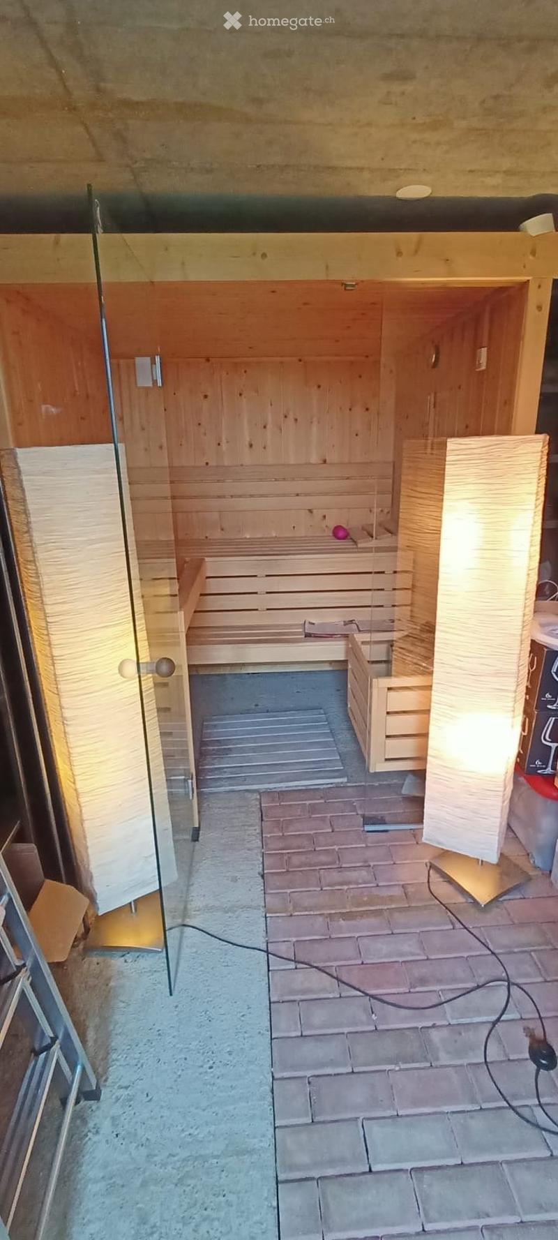 A wooden sauna room with a bench and a small ball placed on it. The room has wooden walls and a glass door leading to a tiled area.