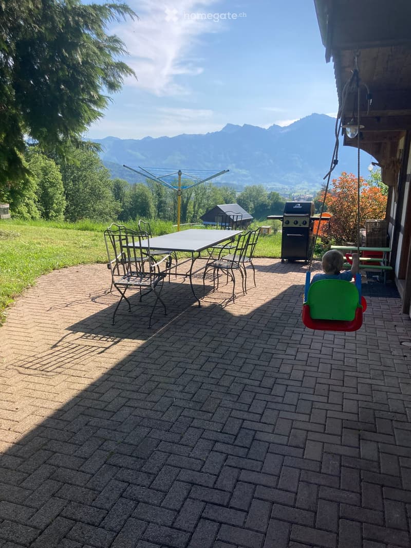 Outdoor patio with brick flooring, a swing, a table with chairs, and a grill, overlooking a scenic mountain view.