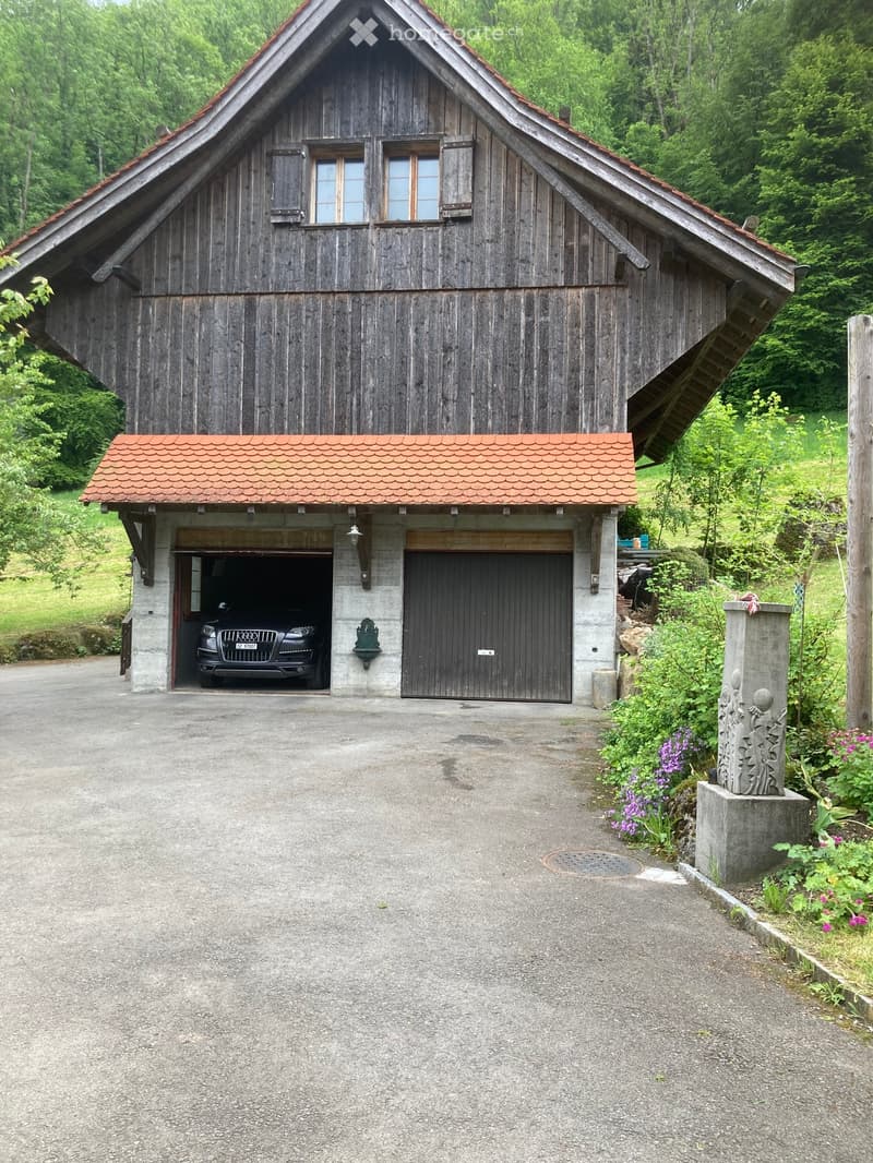 A wooden garage with a sloped roof and two large doors, one open revealing a parked car. The surrounding area includes greenery and a decorative stone pillar.