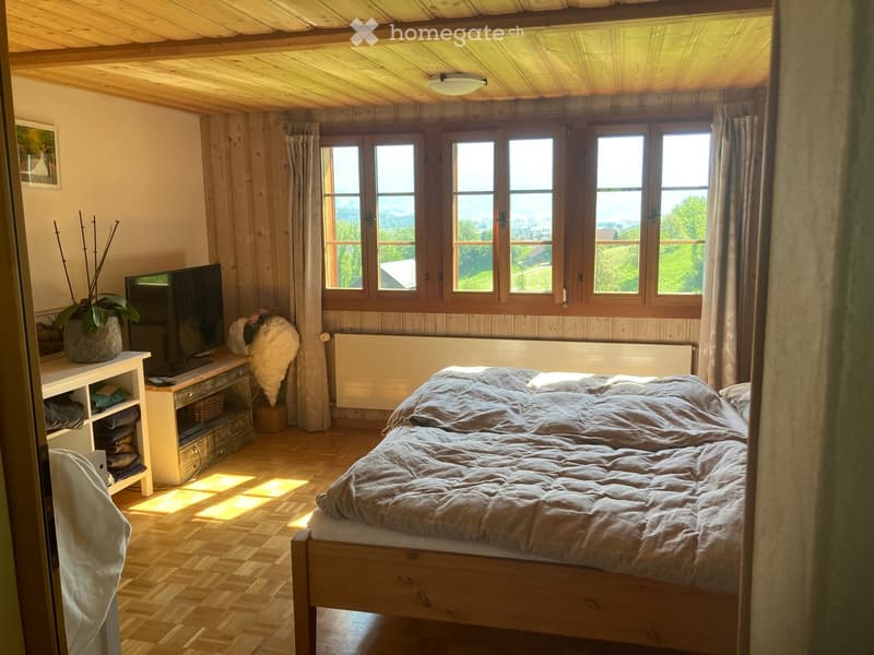 Cozy bedroom with wooden walls and ceiling, large windows with curtains, a bed with a quilt, a nightstand, a chair, a table with a television, and a potted plant.