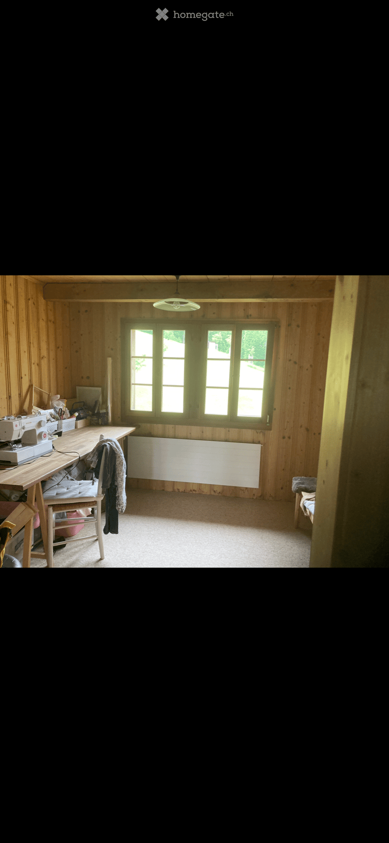 Wooden-walled room with a large window, a sewing machine on a table, a chair, and a radiator.