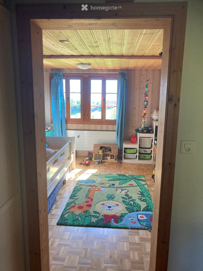 Brightly lit children's bedroom with wooden walls and floors, featuring a large animal-themed rug, a bed, shelves with toys, and a small table with chairs.
