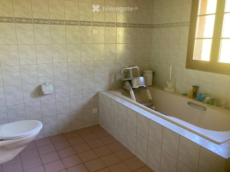 Bathroom with a toilet, bathtub, and a small window letting in natural light. The walls are tiled with a decorative border, and there is a toilet paper holder and a toilet brush visible.
