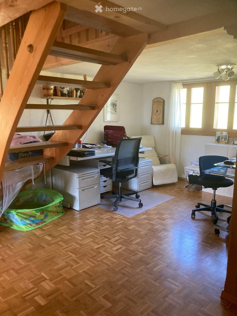A well-lit room with wooden flooring, a staircase leading to an upper level, a desk with a chair, a sofa, a small stool, a printer, and a laptop on a glass table with another chair.