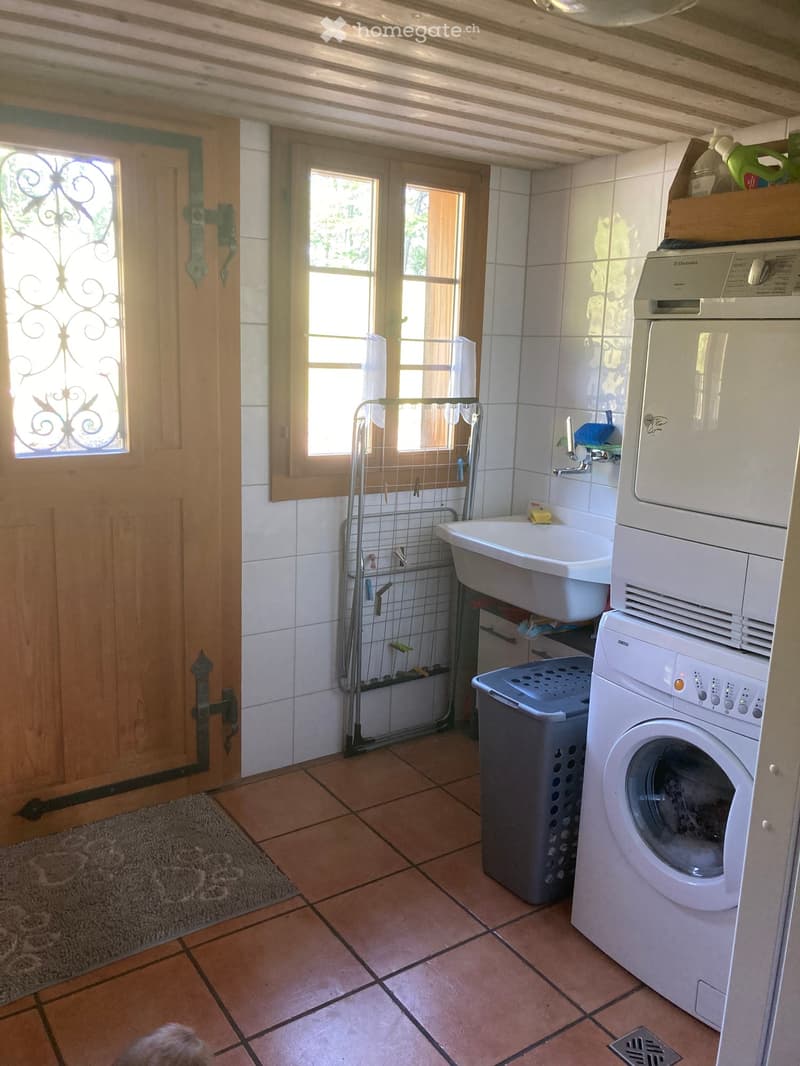A well-lit bathroom with a sink, washing machine, laundry basket, and a drying rack. The room features tiled walls and floors, a wooden door with a decorative glass panel, and a window with a curtain.