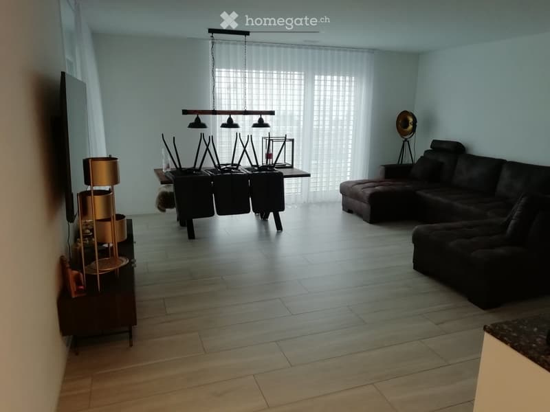 Modern living room with light wooden flooring, large windows with white blinds, a dark brown sectional sofa, a coffee table with chairs, a hanging lamp, and a TV screen on the wall.