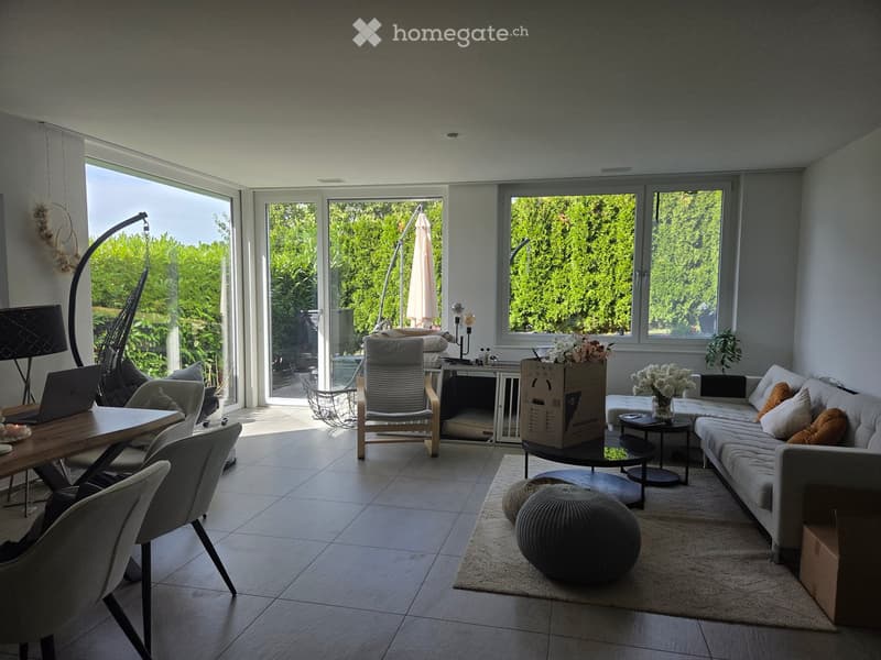 Bright living room with large windows and sliding doors leading to a patio, featuring a grey sofa, coffee table, round stools, dining table with chairs, and decorative vases with flowers.