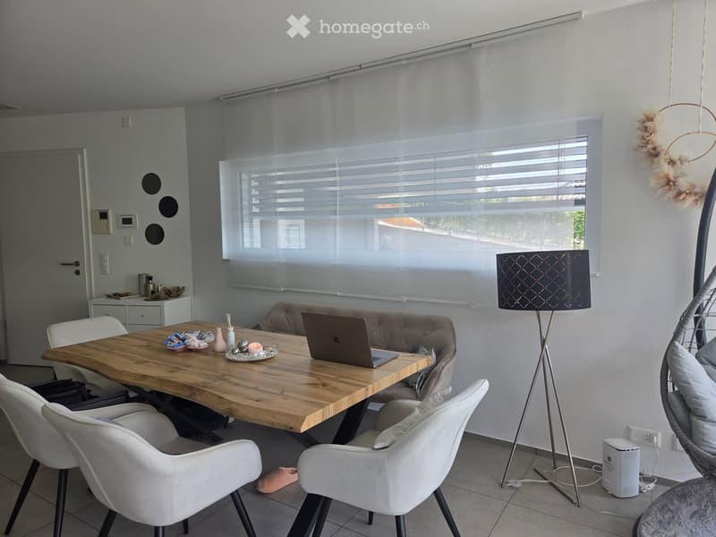 Modern living room with a wooden dining table, white chairs, a laptop, a grey sofa, a floor lamp, and a wicker hanging chair.