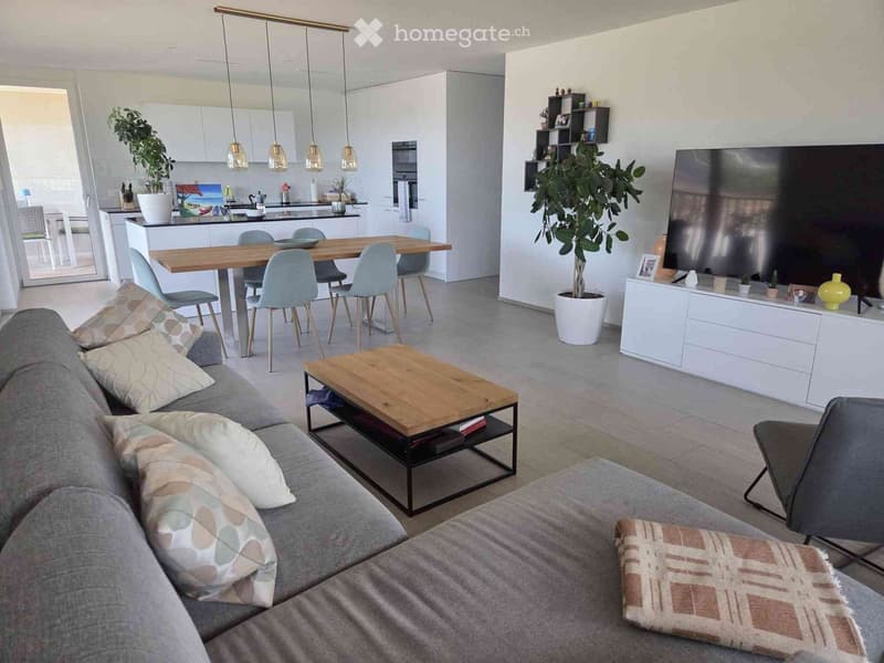 Modern open-plan living and dining area with a grey sofa, wooden coffee table, dining table with chairs, and a large television. The space includes a kitchen area with a white island and pendant lights.