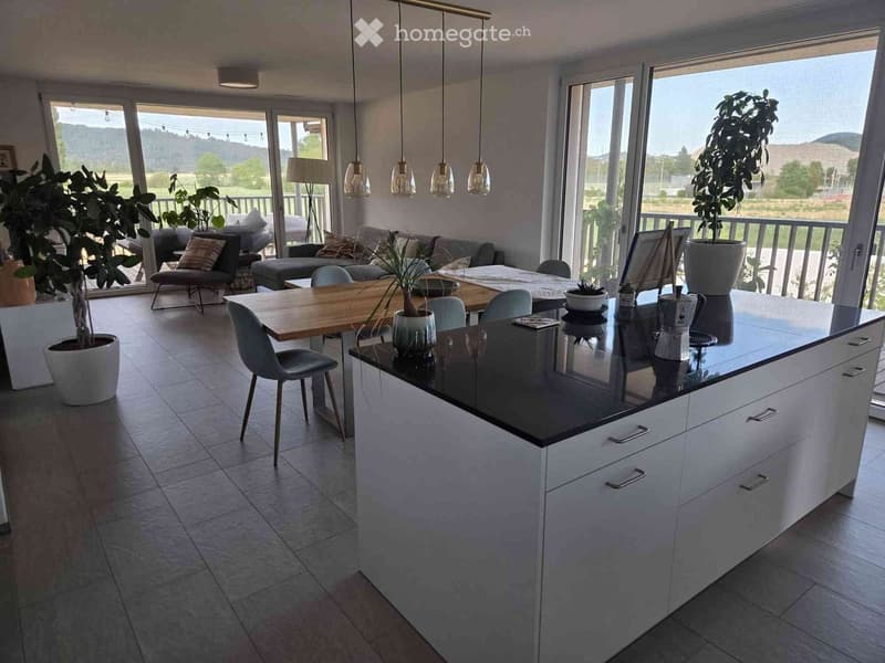 Modern kitchen with a central island, dining area, and living space. Features include pendant lights, large windows with a scenic view, and contemporary furniture.