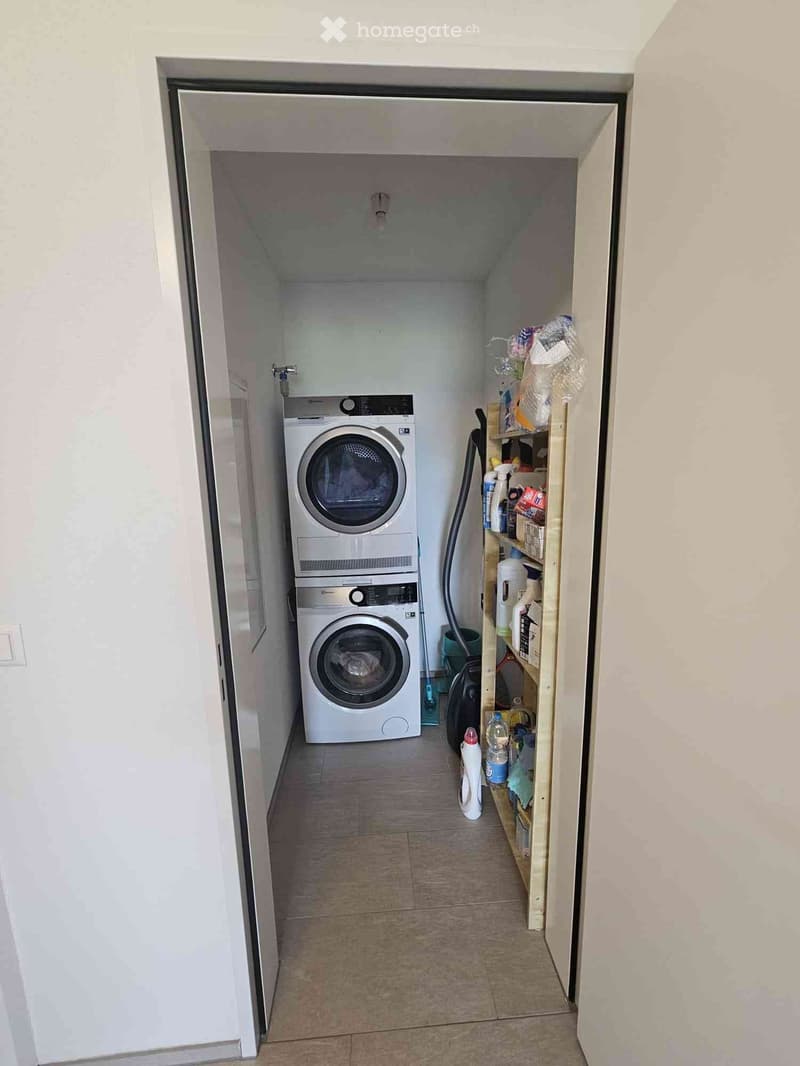 A small, well-lit laundry area with a stacked washer and dryer, a wooden shelf holding cleaning supplies, and a vacuum cleaner nearby.