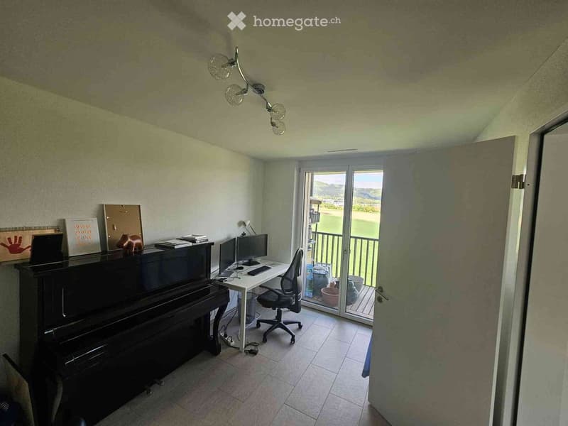 A well-lit room with a piano, desk, chair, laptop, and a view through a sliding glass door.