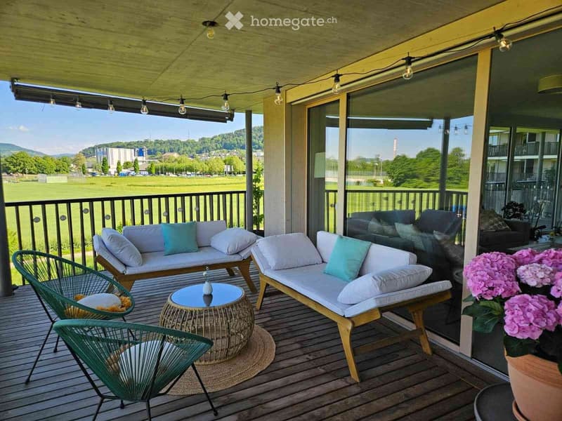 A wooden balcony with a view of a green field and mountains, furnished with a sofa, chairs, a small table, and potted flowers.