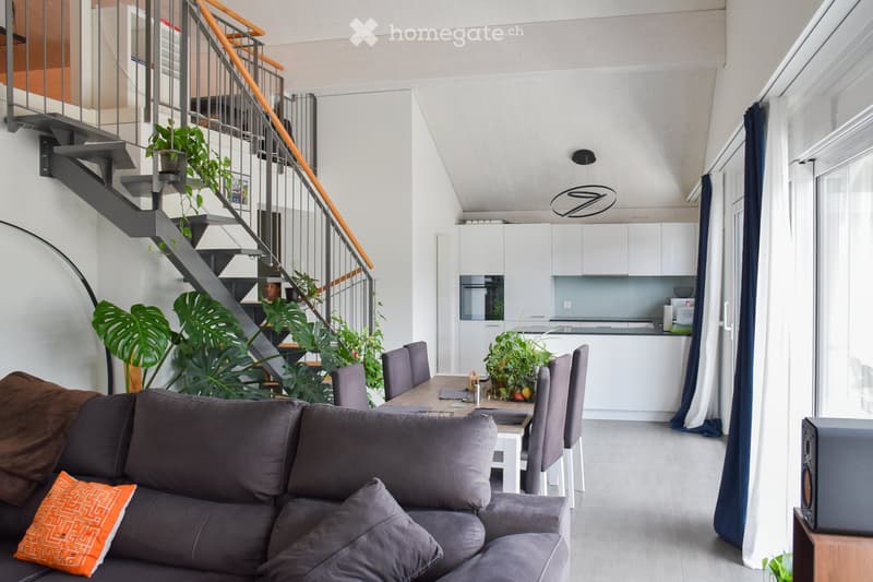 Open-plan living and dining area with a grey sectional sofa, wooden dining table, modern kitchen, and staircase leading to an upper level.