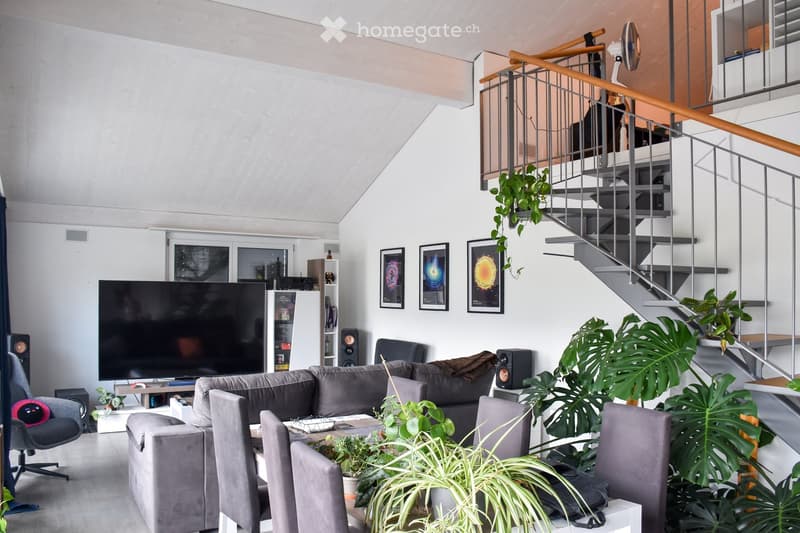 Modern living room with grey sectional sofa, coffee table, large flat-screen TV, speakers, and several potted plants. The room features a staircase with a wooden railing and a sleek, contemporary design.