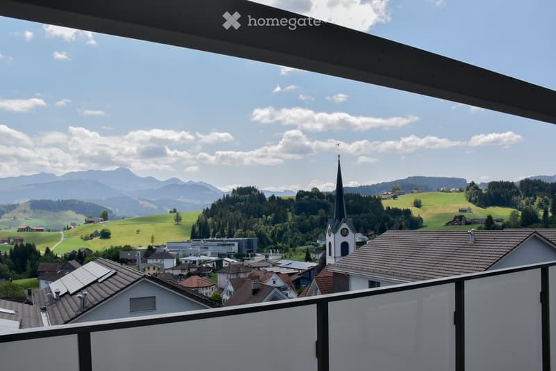Balcony overlooking a picturesque village with a church tower, surrounded by green hills and distant mountains under a partly cloudy sky.