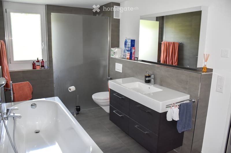 Modern bathroom with a bathtub, sink, toilet, large mirror, and various toiletries on the counter. The space features dark grey tiles and a sleek design.