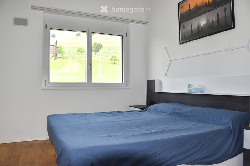 Modern bedroom with a blue bedspread, wooden floor, large windows overlooking a green field, and a mounted picture frame on the wall.