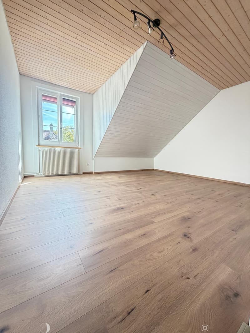 Empty attic room with wooden flooring, white walls, and a slanted wooden ceiling. Features a window with a view and a radiator below it.