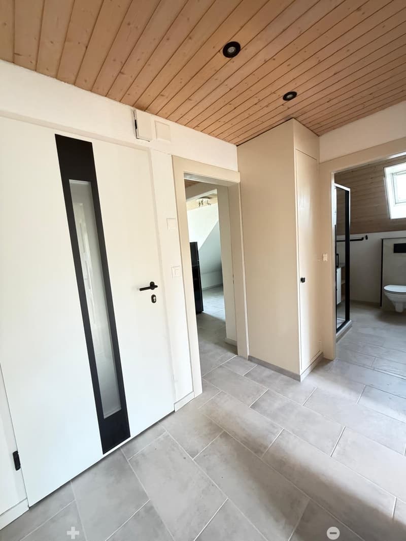 Empty hallway with tiled floor, white walls, and wooden ceiling. Features a door leading to a bathroom with a visible toilet.