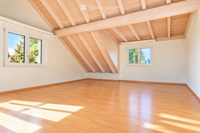 Bright, empty attic room with wooden floors, white walls, and exposed wooden beams on the ceiling. Two large windows offer natural light and a view of greenery outside.