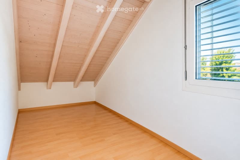 Empty room with wooden flooring, white walls, and a slanted wooden ceiling. A large window with blinds allows natural light to enter.