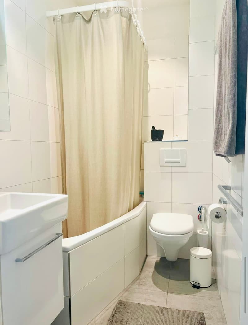 Modern bathroom with white tiled walls and floor, featuring a bathtub, toilet, sink, and a shower area with a curtain. Includes a towel, toilet paper holder, and a small trash bin.