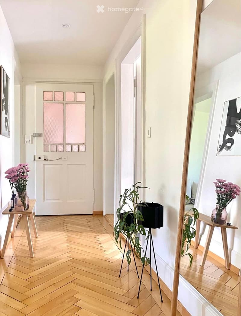 A modern hallway with herringbone wooden flooring, white walls, and a white door with frosted glass panels. There are small tables with vases of pink flowers and a potted plant on stands.