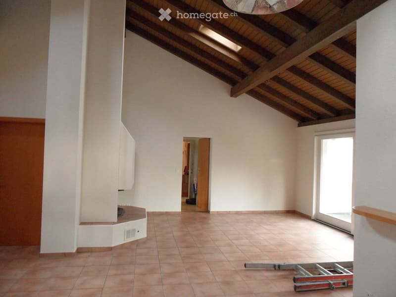 Empty room with exposed wooden beams on the ceiling, tiled floor, and a large window letting in natural light. There is a doorway leading to another room and some construction equipment visible.