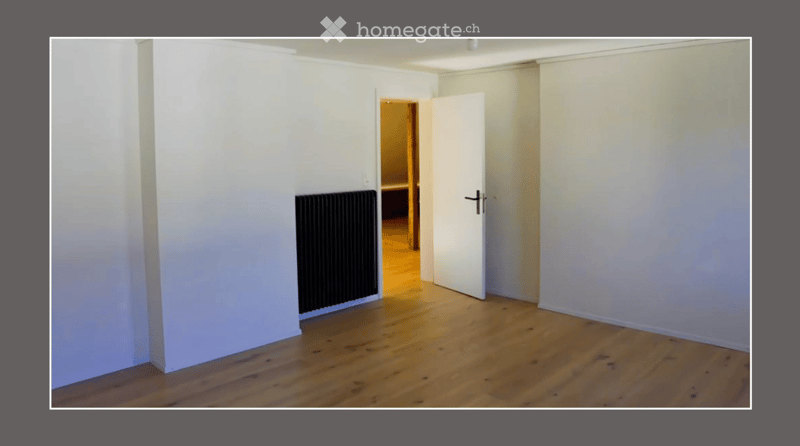 Empty room with light wooden flooring, white walls, and a radiator on one wall. An open door leads to another room with warm lighting.
