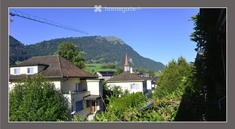 View of a residential area with houses, surrounded by lush greenery and a mountainous backdrop under a clear blue sky.