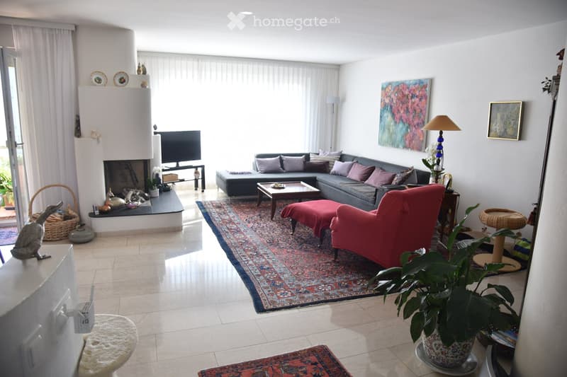 Bright living room with large windows, grey sectional sofa, red armchair, coffee table, fireplace, TV, floor lamp, and decorative items.