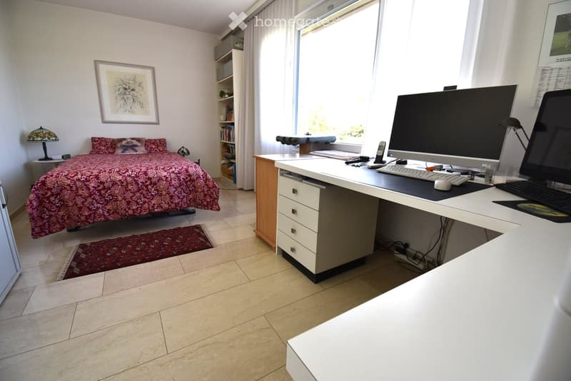 Bright bedroom with a large bed, a desk with a computer setup, a bookshelf, and a lamp. The room features large windows with white curtains and a tiled floor.