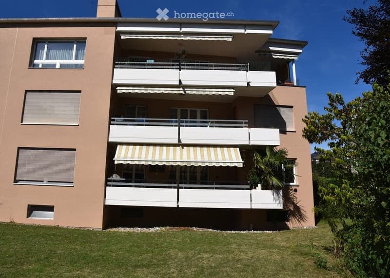 A three-story residential building with beige walls, multiple balconies, striped awnings, and large windows. The building is surrounded by greenery and a well-maintained lawn.
