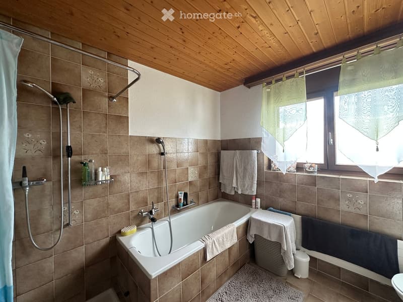 Bathroom with a bathtub, shower, sink, toilet, towels, and a window with a curtain. Wooden ceiling and tiled walls.
