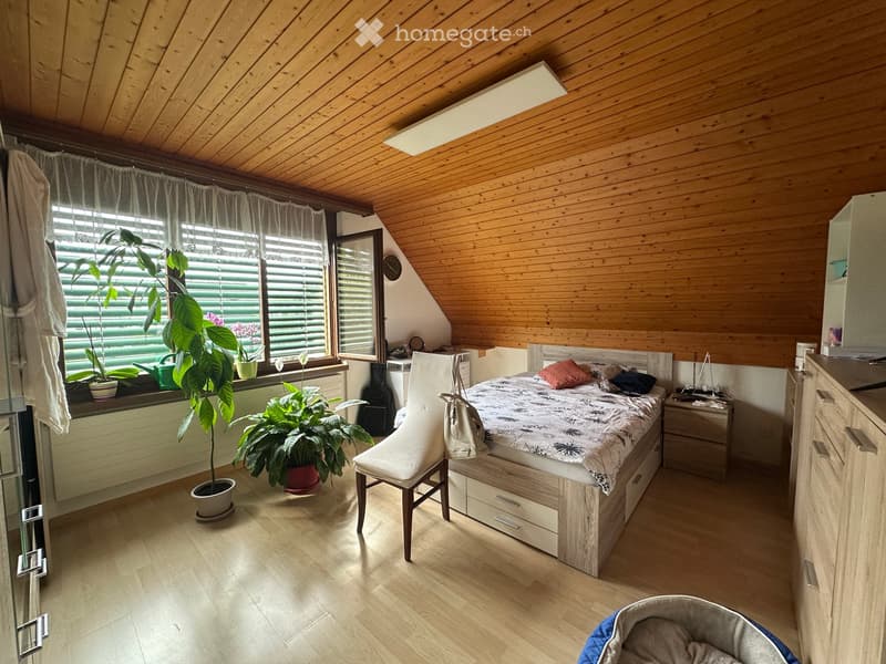 Cozy attic bedroom with wooden walls and ceiling, large windows with blinds, bed with patterned bedding, chair, nightstand, and several potted plants.