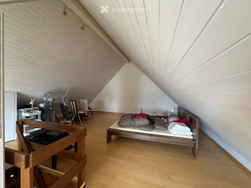 Attic bedroom with wooden floors, slanted ceilings, and a bed with pillows and blankets. Includes a chair, table, and fan.