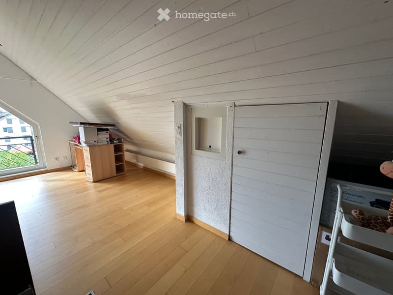 Spacious attic room with wooden floors, slanted ceilings, and large windows. Contains a small wooden cabinet, a desk with books, and a toy giraffe.