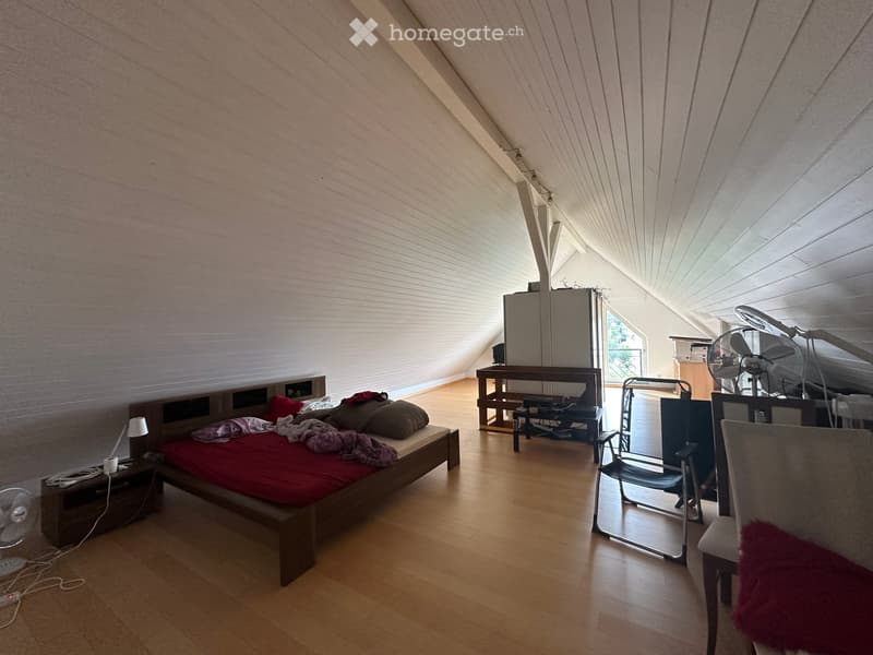Bright attic bedroom with wooden floors, slanted ceilings, and large windows. Features a bed with red bedding, a nightstand, a fan, a chair, a small table, and a mirror.