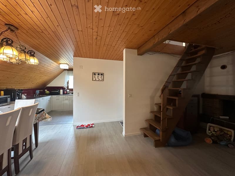 Open-plan living and dining area with wooden ceiling beams, staircase leading to upper level, modern kitchen area with white cabinets and stainless steel appliances, dining table with chairs, and a cozy fireplace area.