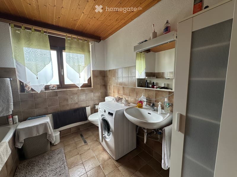 Bathroom with a sink, toilet, washing machine, bathtub, mirror, and various toiletries. The room features tiled walls and a wooden ceiling.
