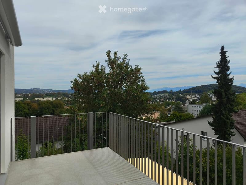 Balcony with metal railing, overlooking a cityscape with trees, buildings, and distant mountains under a partly cloudy sky.