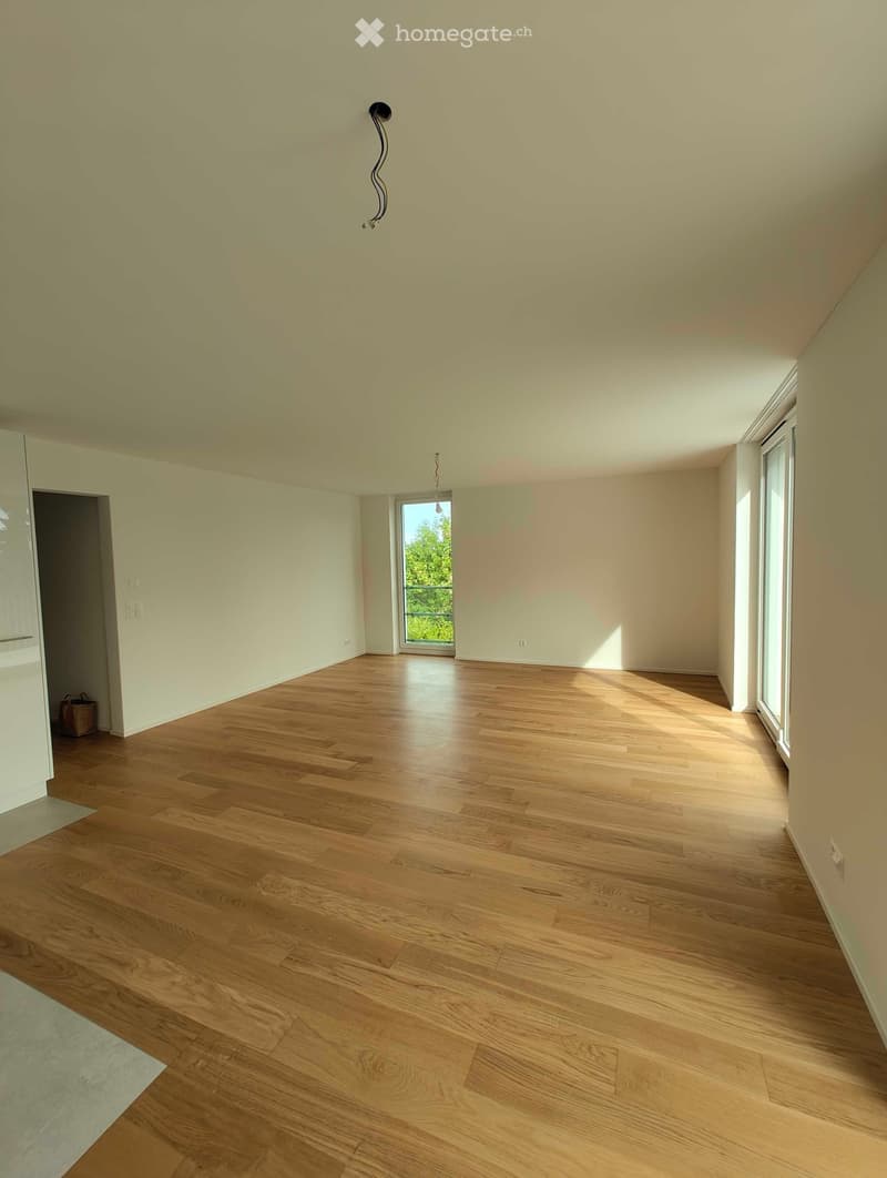 Empty room with light-colored walls, wooden flooring, and large windows allowing natural light to enter. A small bag is placed near the doorway.